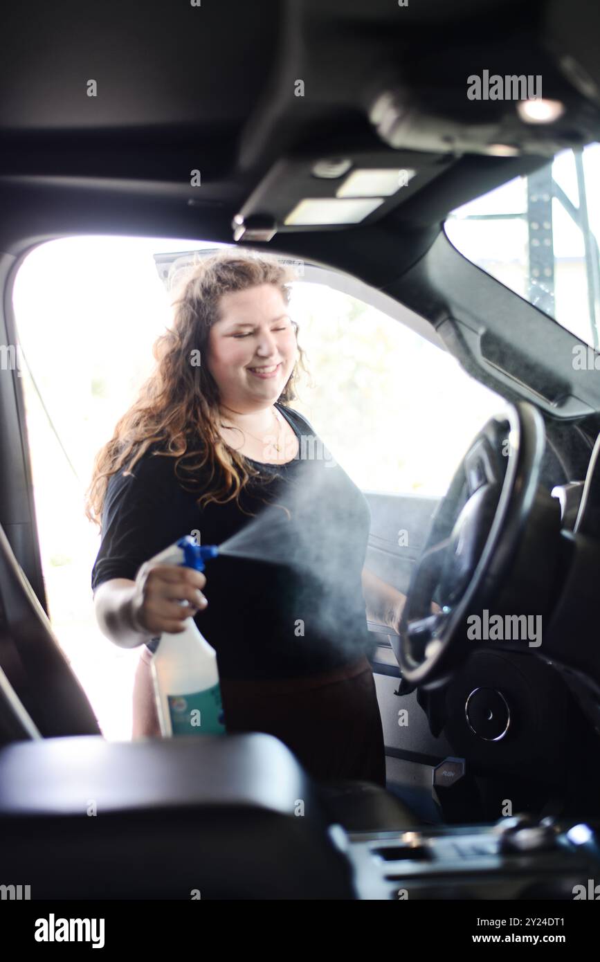 Femme pulvérisant la solution de nettoyage à l'intérieur d'une voiture, souriant tout en cle Banque D'Images