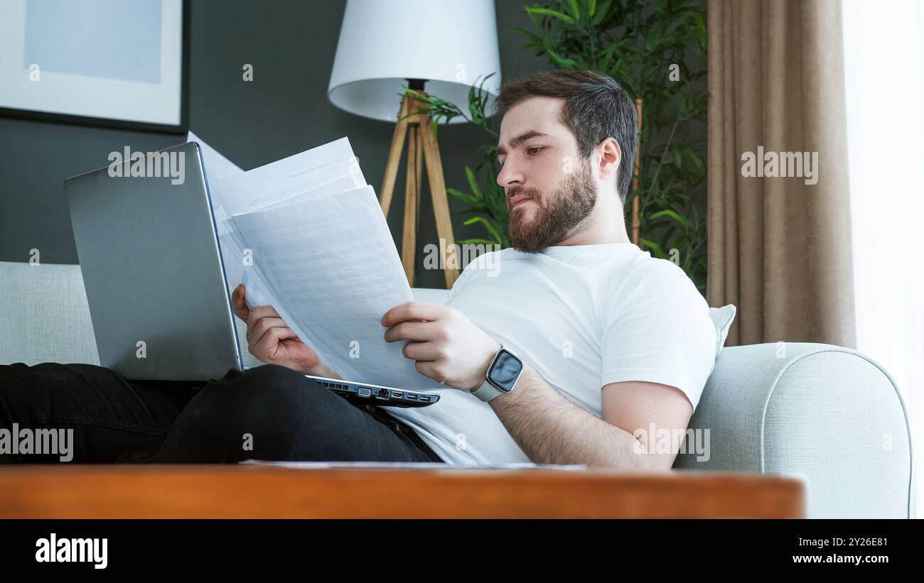 Un homme allongé sur un canapé examine des documents financiers tout en utilisant un ordinateur portable. Concentration et responsabilité impliquées dans la gestion des finances personnelles, des factures et Banque D'Images