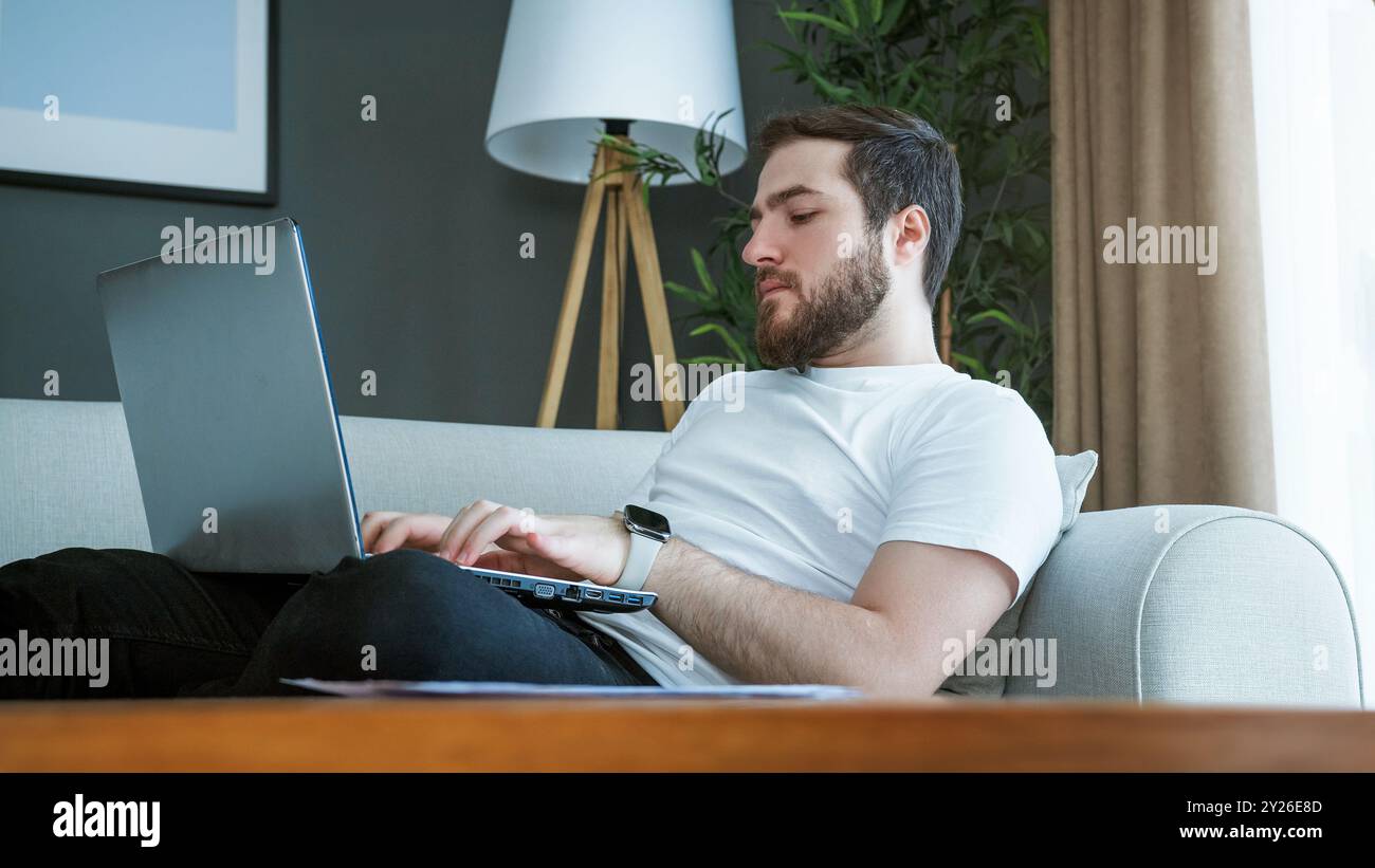 Un homme allongé sur un canapé travaillant sur un ordinateur portable. Concentration et responsabilité impliquées dans la gestion des finances personnelles, des factures et des documents importants. Banque D'Images