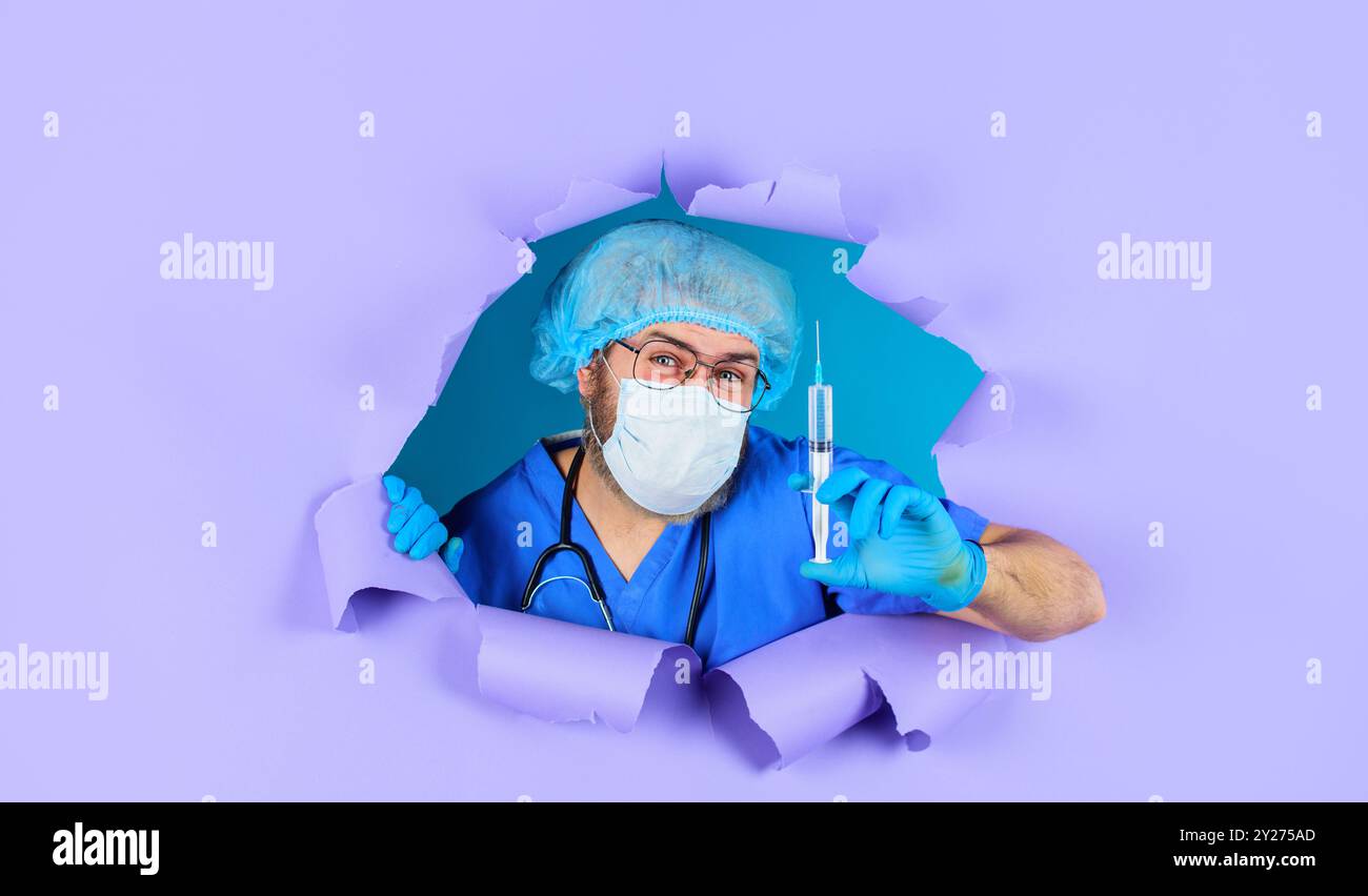 Vaccination des personnes. Docteur en masque médical et capuchon chirurgical avec seringue regardant à travers le trou dans le papier. Infirmière souriante ou médecin en uniforme et Banque D'Images