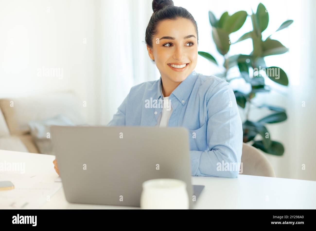 Photo d'une belle femme d'affaires arabe ou indienne positive à succès, chef de projet, pdg de l'entreprise, est assise à un bureau, travaille sur un ordinateur portable, la stratégie de planification et la promotion, regarde loin et sourit Banque D'Images