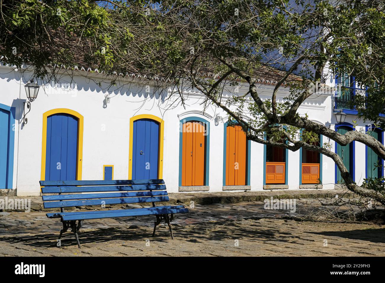 Vue sur les façades de maisons coloniales avec des portes en bois colorées au soleil avec un banc bleu et des branches d'arbres en face, ville historique Paraty, Brésil, UNES Banque D'Images