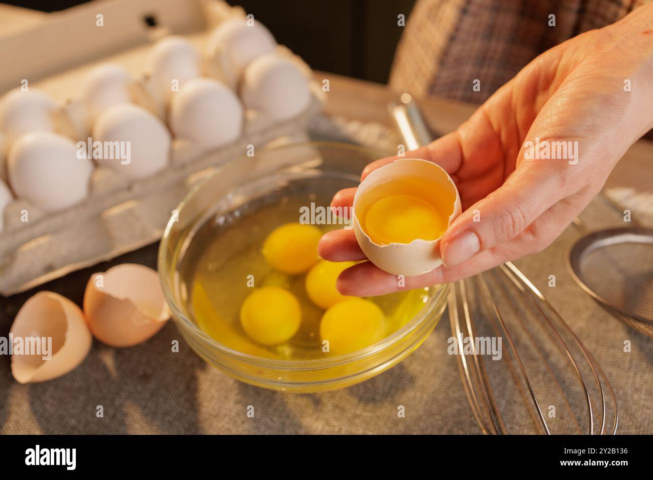 La main d'une femme tenant un œuf de poule cassé gros plan sur un bol d'œufs crus. Il y a plusieurs jaunes d'oeufs dans le bol et plus d'oeufs dans une boîte en carton Banque D'Images