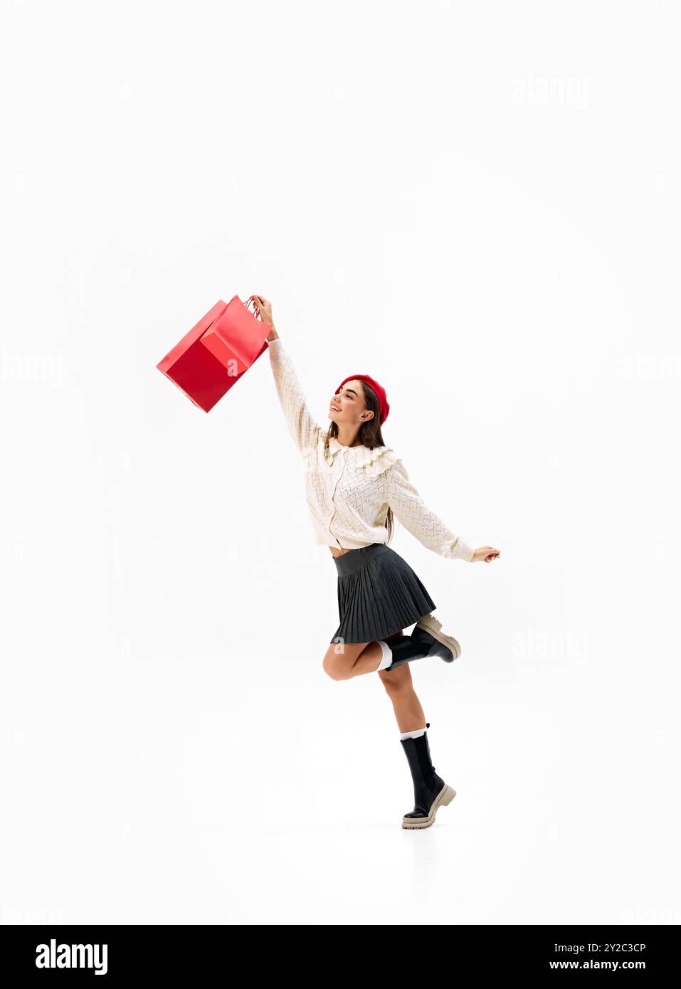 Jeune femme saute joyeusement dans l'air, tenant des sacs à provisions rouges vibrants sur fond blanc de studio. Une virée shopping réussie Banque D'Images