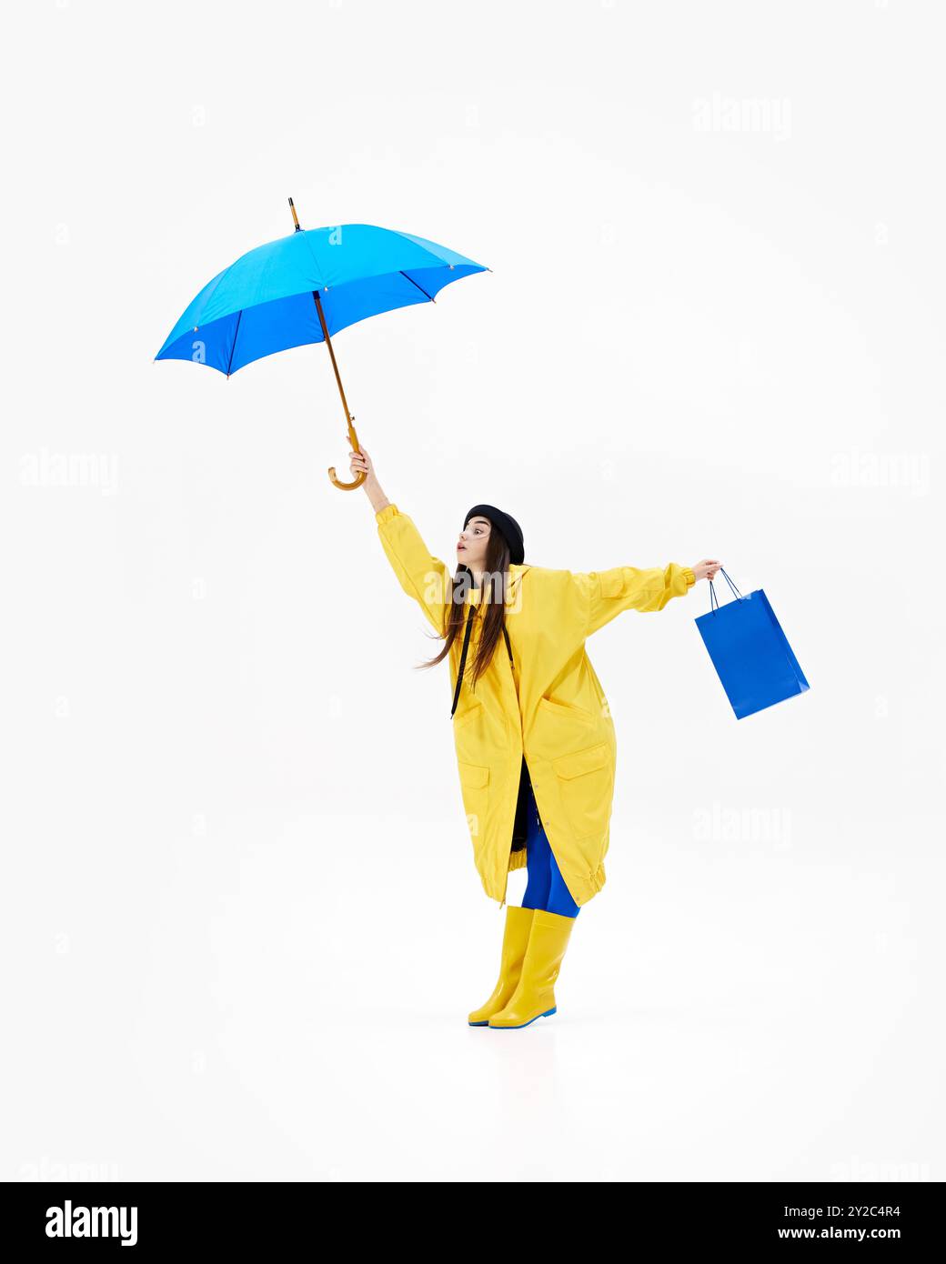 Femme joyeuse dans l'imperméable vibrant saute avec sac à provisions bleu et parapluie, embrassant le temps pluvieux sur fond blanc. Banque D'Images