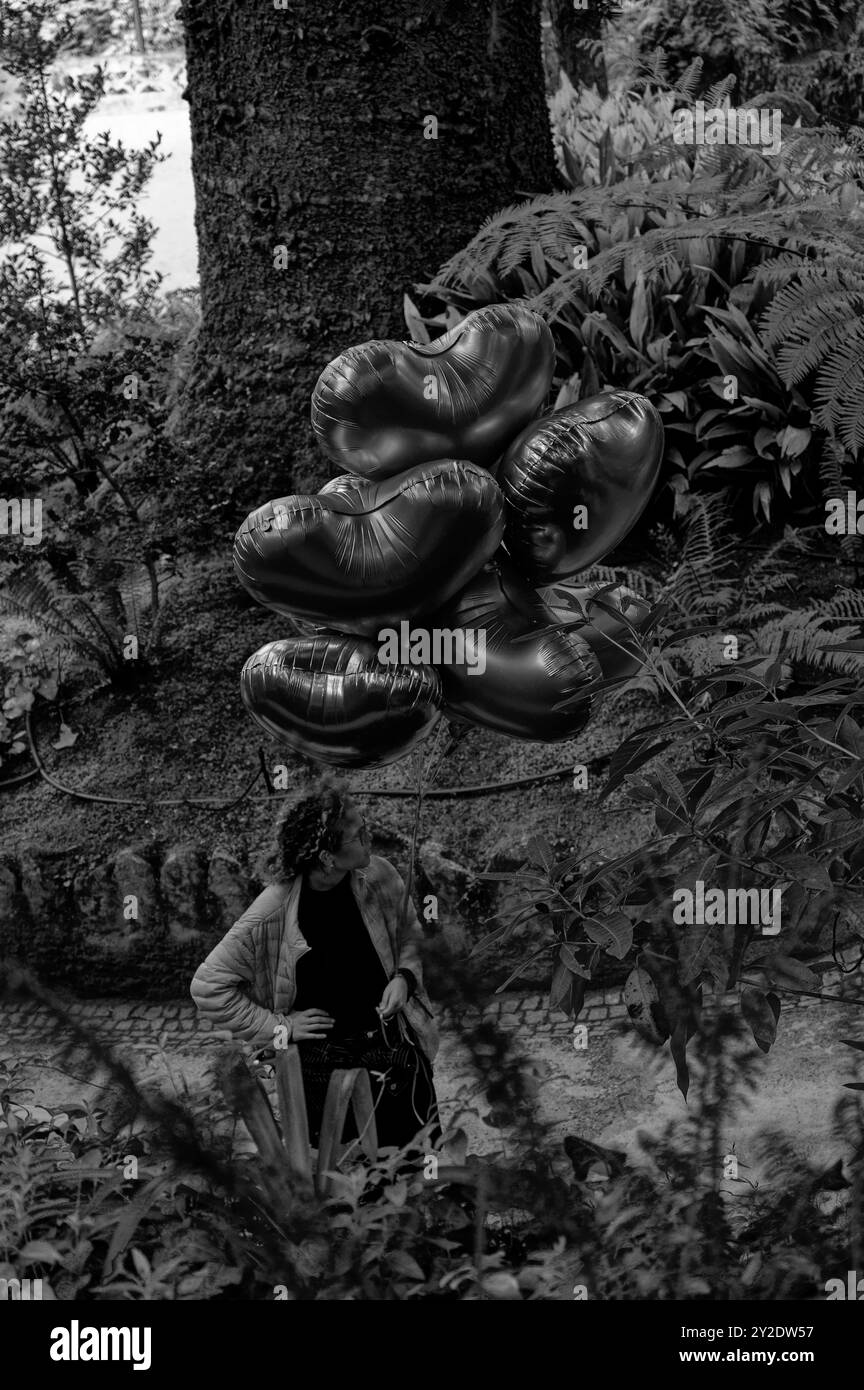 Monochrome d'une femme tenant un groupe de ballons en forme de cœur, flottant contre un jardin luxuriant en toile de fond à Quinta da Regaleira Banque D'Images