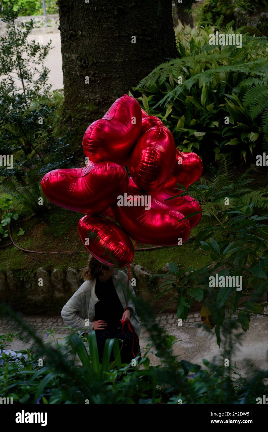 Un groupe de ballons rouges vibrants en forme de cœur flotte sur un jardin luxuriant en toile de fond à Quinta da Regaleira Banque D'Images