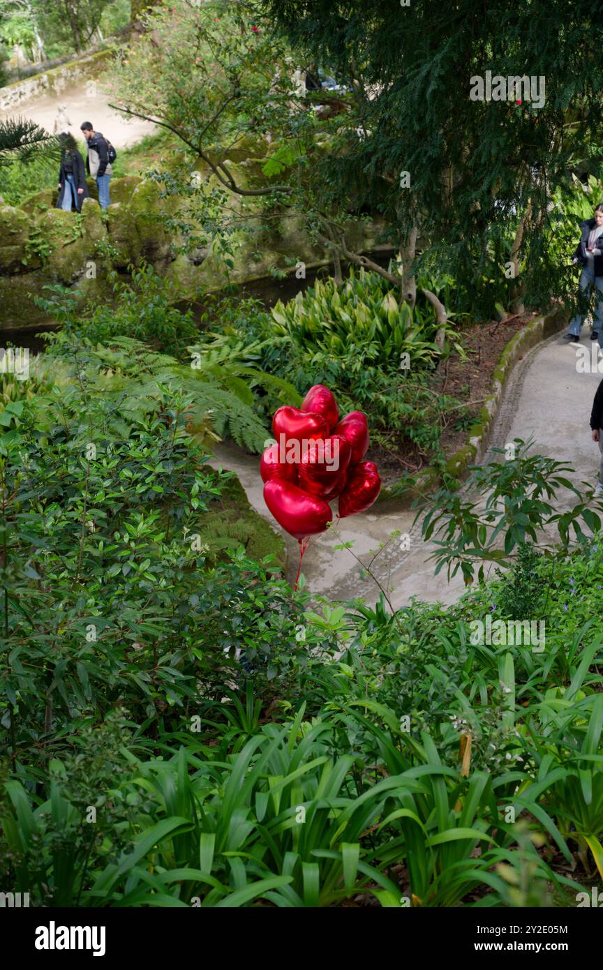 Un groupe de ballons rouges vibrants en forme de cœur flotte doucement au milieu de la riche verdure des sentiers de jardin de Quinta da Regaleira Banque D'Images