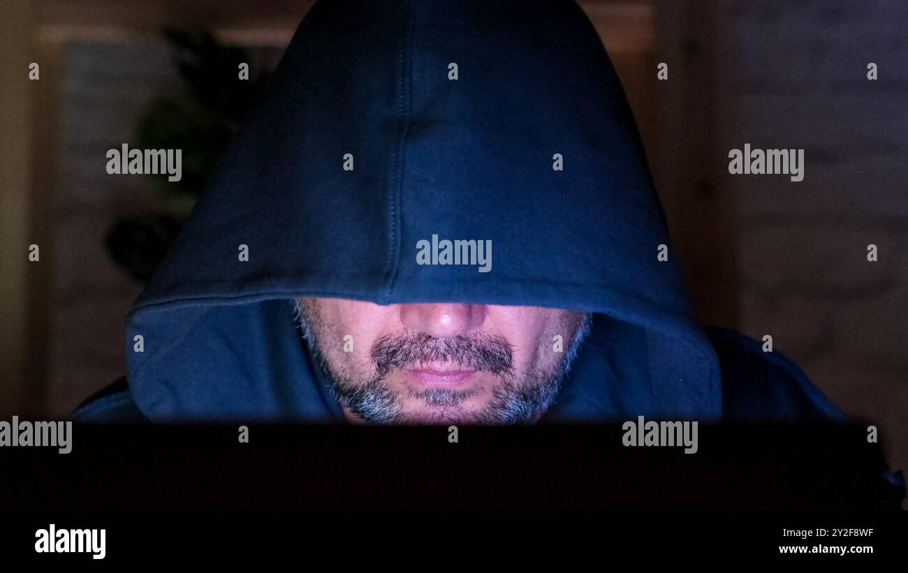 Pirate à capuche d'âge moyen barbu sur un ordinateur portable la nuit de gros plan. Homme utilisant un ordinateur travaillant à la maison Banque D'Images