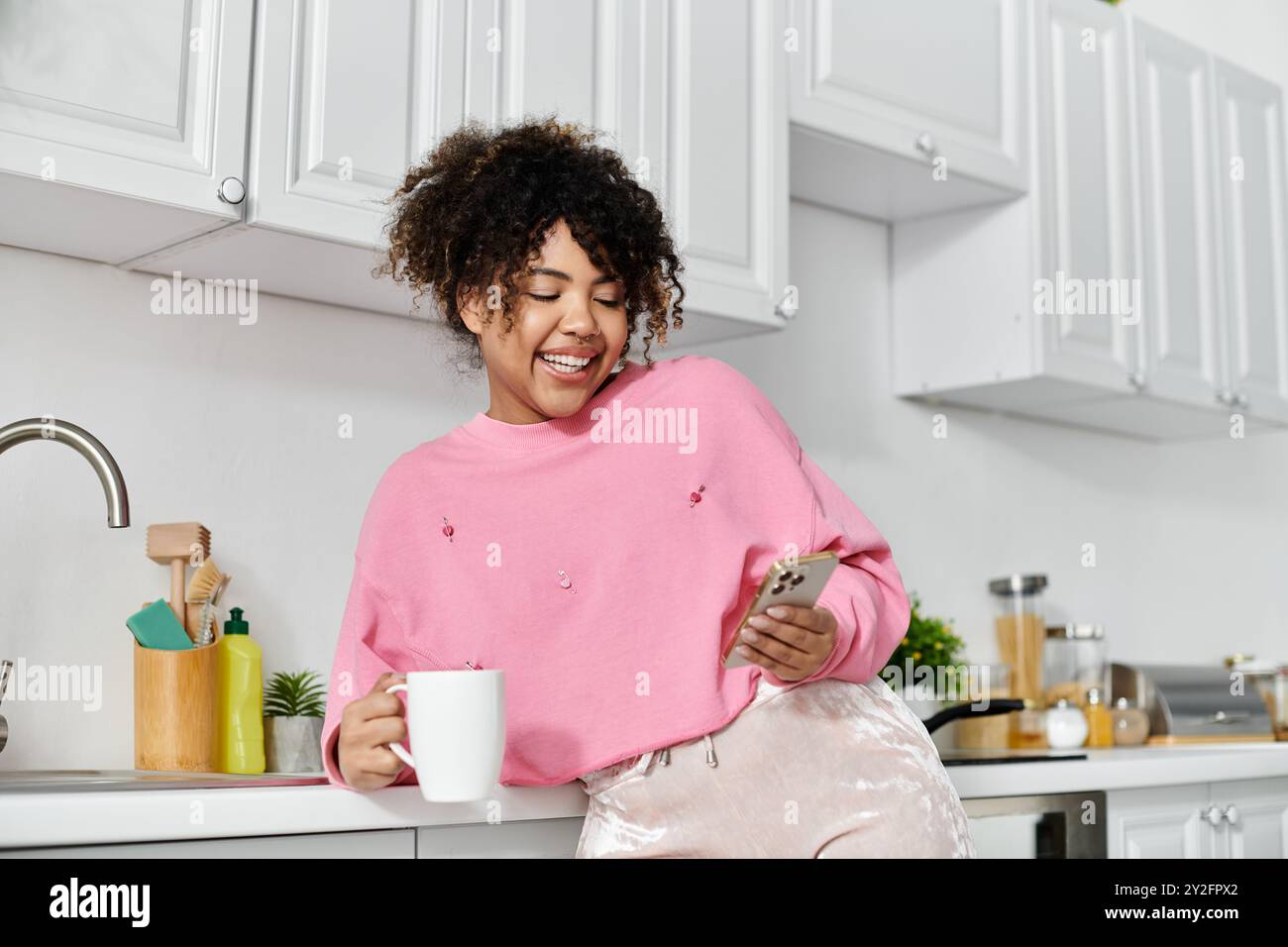 Femme souriante se détend avec un café tout en utilisant son téléphone dans une cuisine lumineuse. Banque D'Images