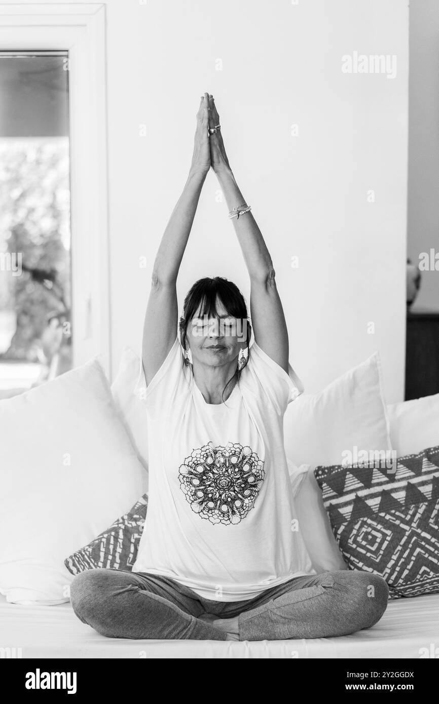Femme mi-adulte méditant sur son canapé à la maison pendant la journée, avec la lumière naturelle et les paumes pressées ensemble au-dessus de sa tête. Banque D'Images