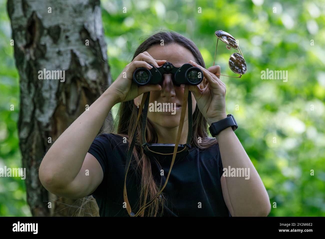 Une femme tient des jumelles sur ses yeux, observant attentivement la faune dans une forêt verte vibrante, entourée d'arbres et de feuillage naturel par une journée ensoleillée. Banque D'Images