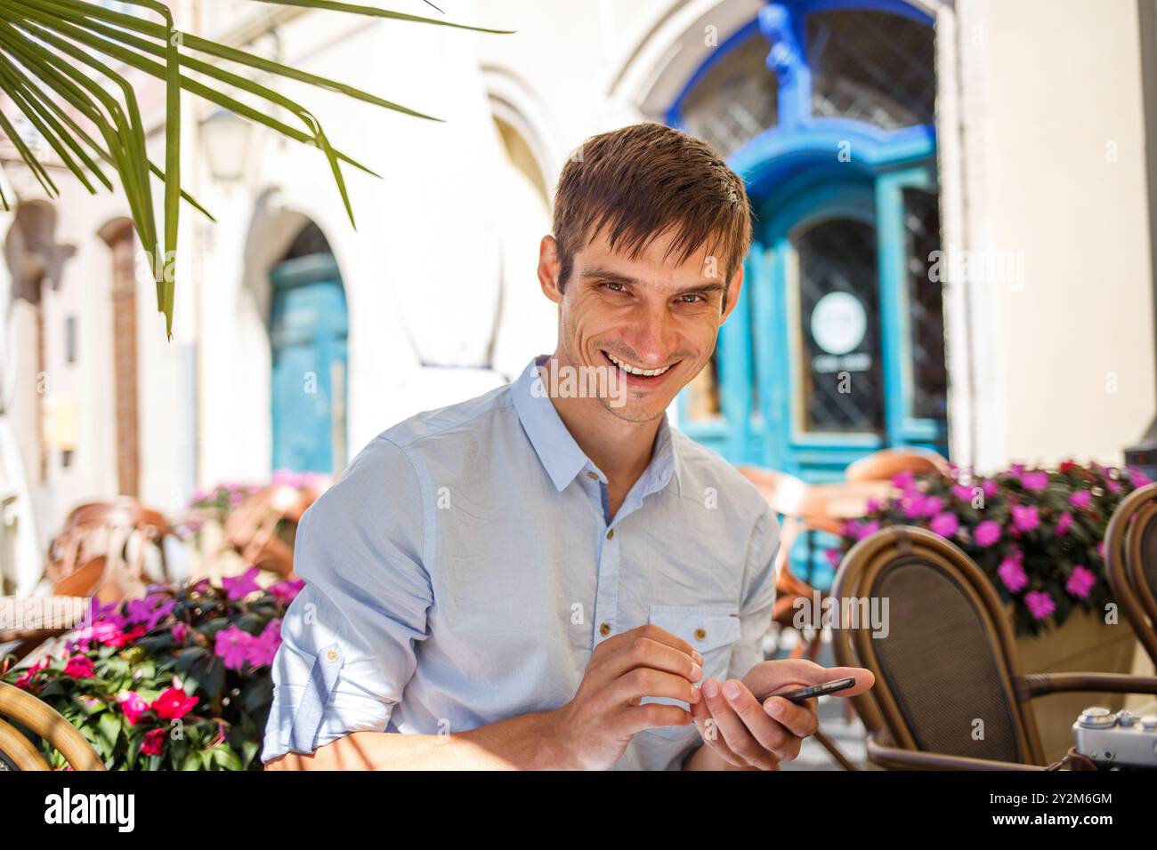 Homme souriant tout en utilisant un smartphone dans un café extérieur ensoleillé au printemps. Banque D'Images