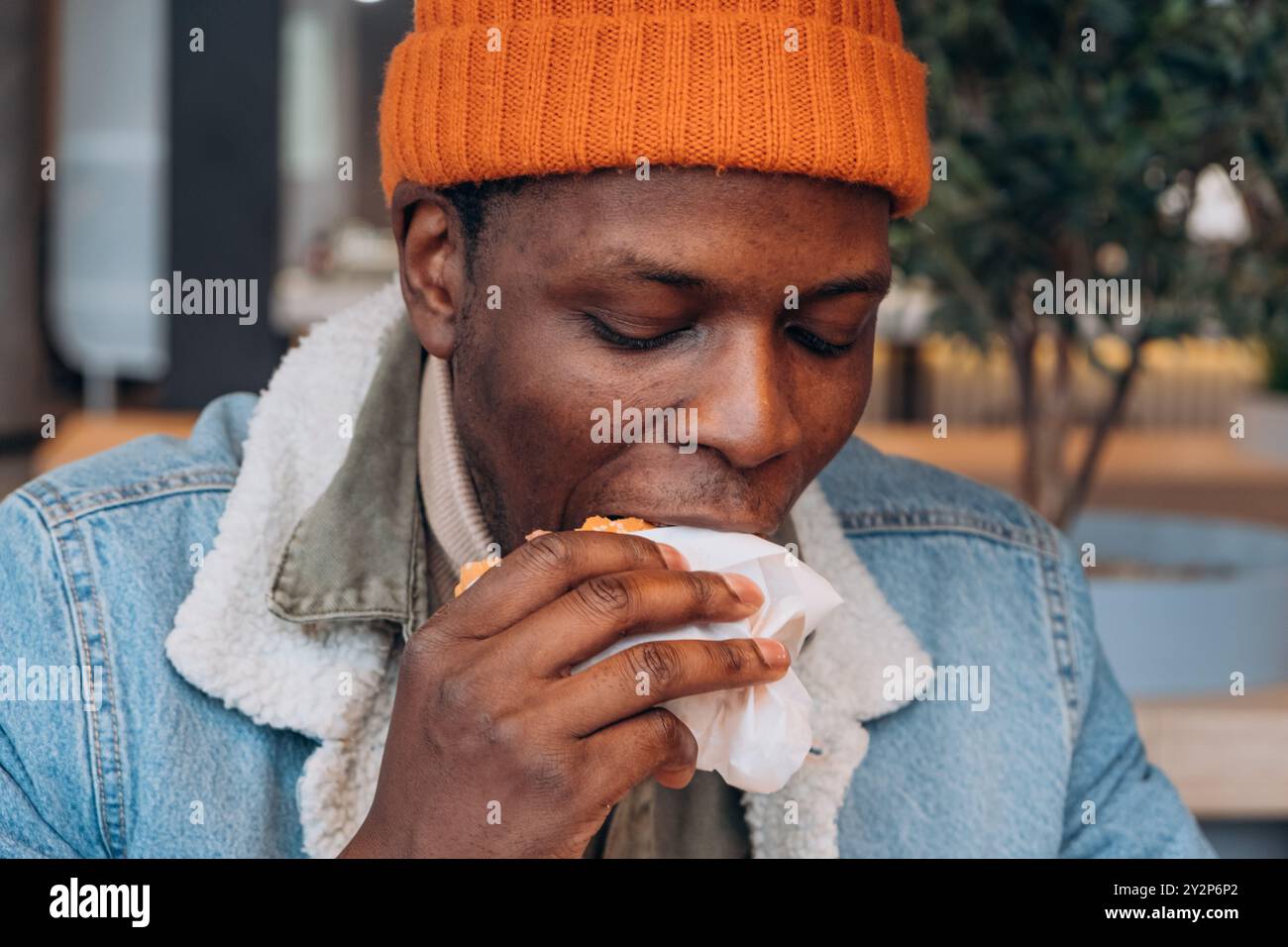 Homme afro-américain mangeant hamburger en bonnet orange et veste en denim dans un environnement extérieur décontracté. Personne affamée dévorant la restauration rapide à l'aire de restauration de rue avec les yeux rabaissés de la hâte. Banque D'Images