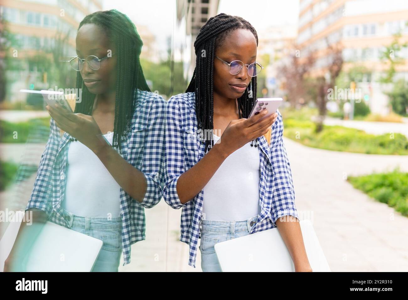 Une femme noire concentrée gère son emploi du temps chargé en utilisant un smartphone et en transportant un ordinateur portable dans un quartier d'affaires urbain Banque D'Images