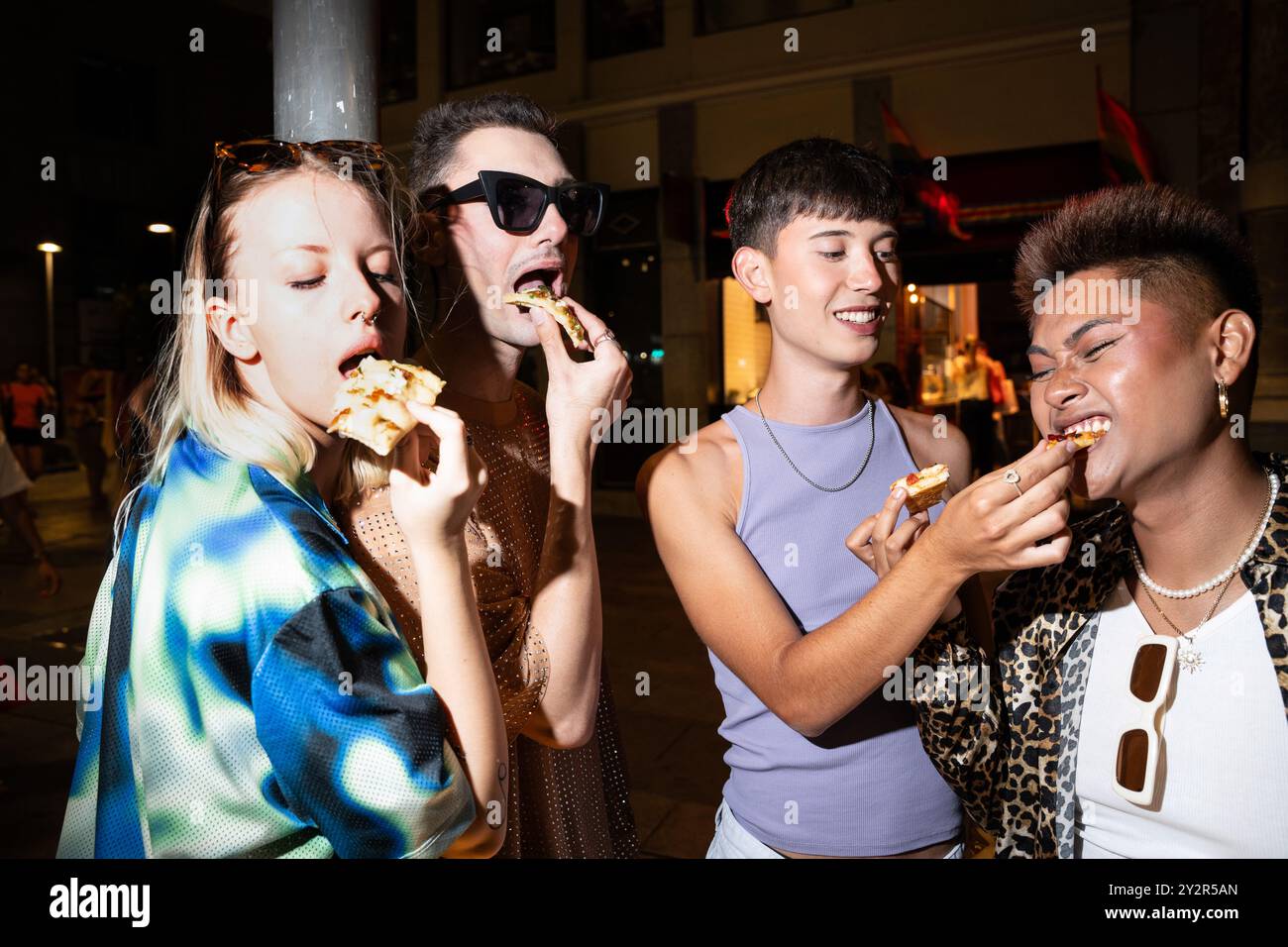 Groupe d’amis LGBTQIA+ partageant un moment joyeux tout en mangeant de la nourriture de rue pendant une soirée, mettant en valeur la diversité et l’amitié. Banque D'Images