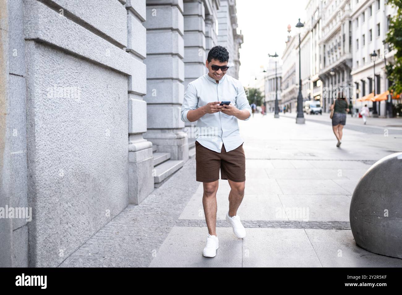 Un jeune homme latin vêtu d'une chemise blanche décontractée et d'un short marron sourit alors qu'il utilise son smartphone tout en marchant dans une rue animée de la ville. Banque D'Images