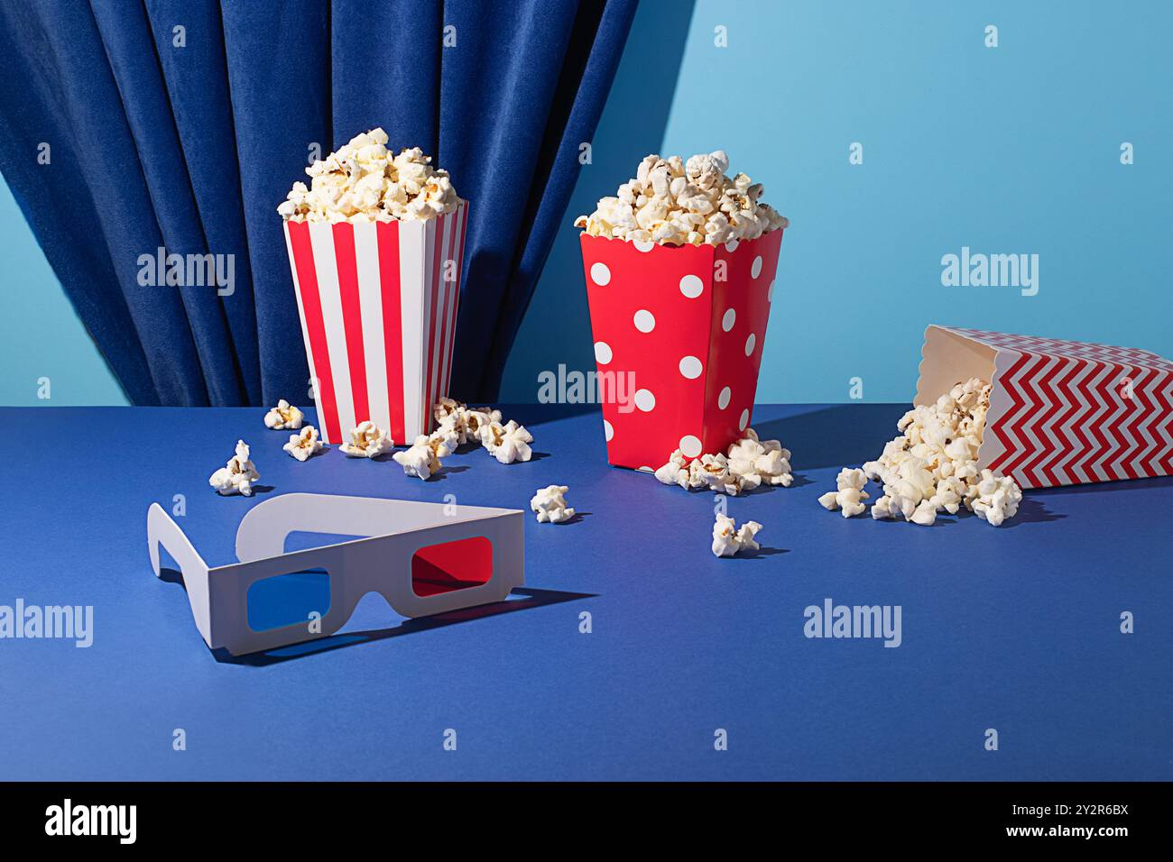 Seaux à pop-corn colorés et lunettes 3D placés sur un fond bleu vif et rideau bleu drapé, capturant l'essence d'une soirée cinéma amusante. Banque D'Images