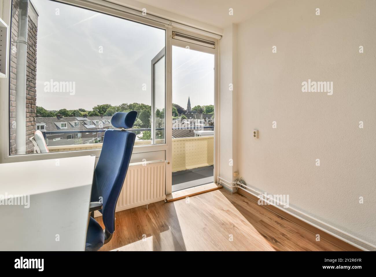 Une pièce inoccupée avec un sol stratifié, une fenêtre ouverte et une chaise de bureau bleue unique. La lumière du jour remplit l'espace, offrant une vue sur les toits. Banque D'Images