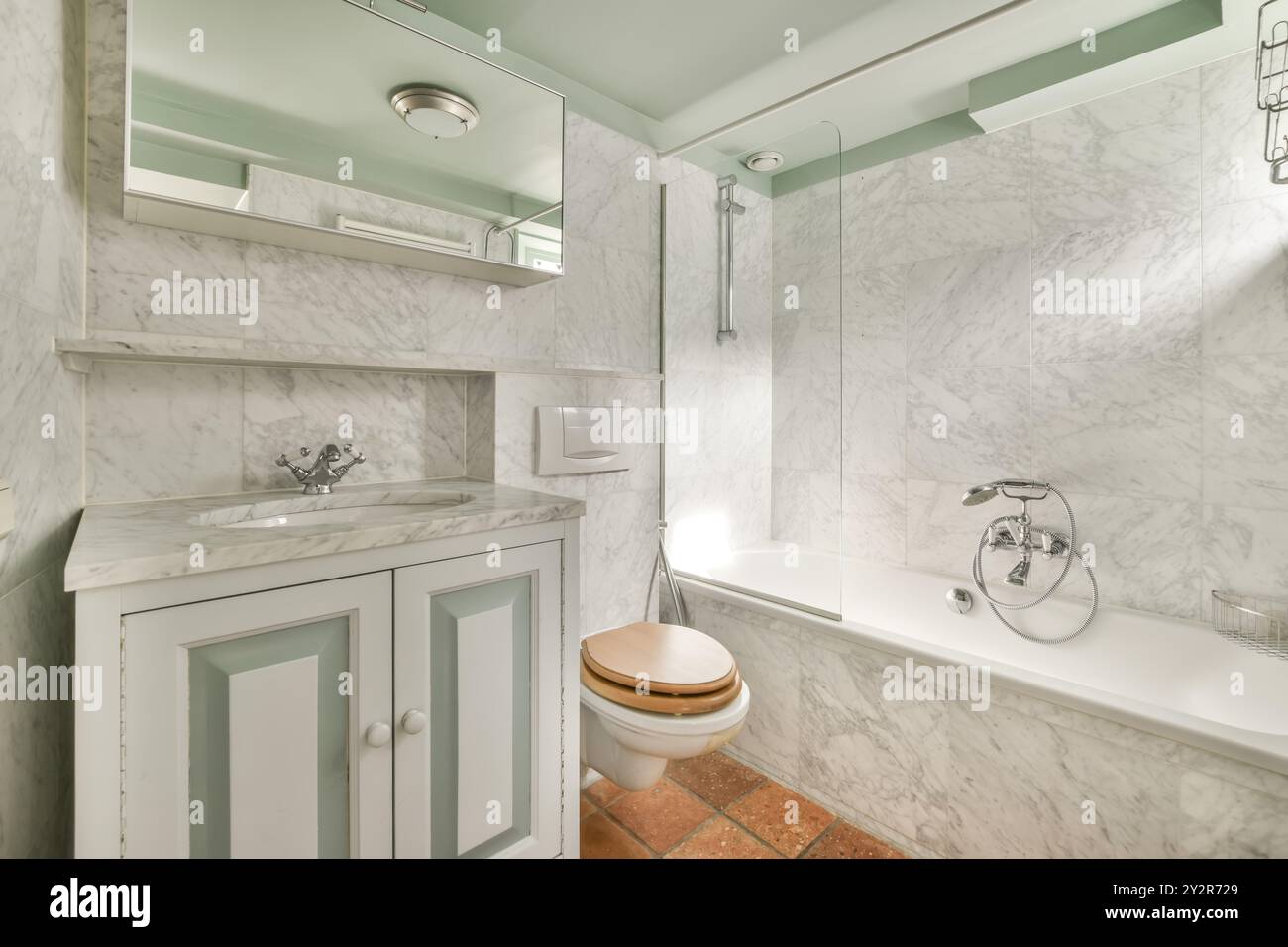 Cette salle de bains décorée avec goût comprend des murs en marbre, une baignoire avec pomme de douche, une armoire à miroirs et un meuble de toilette en bois. Banque D'Images