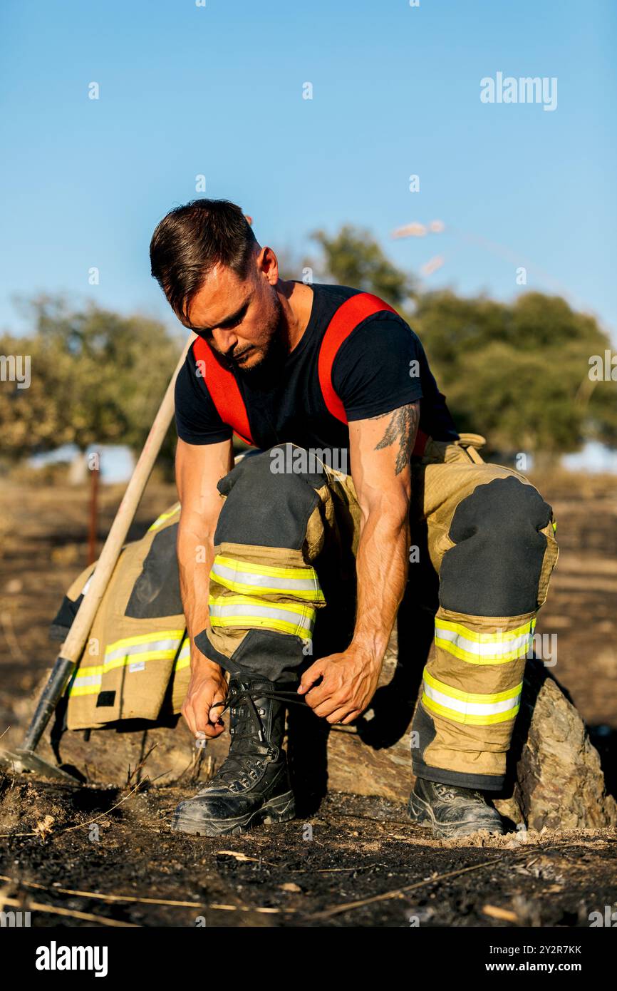 Un pompier concentré ajuste ses bottes de protection et son équipement au milieu d'un environnement forestier, reflétant la préparation et le sérieux requis pour les sapins sauvages Banque D'Images