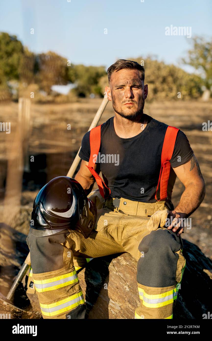 Un pompier déterminé s'assoit calmement après avoir combattu un incendie de forêt, tenant un casque et s'appuyant sur un rocher au milieu d'un paysage carbonisé Banque D'Images
