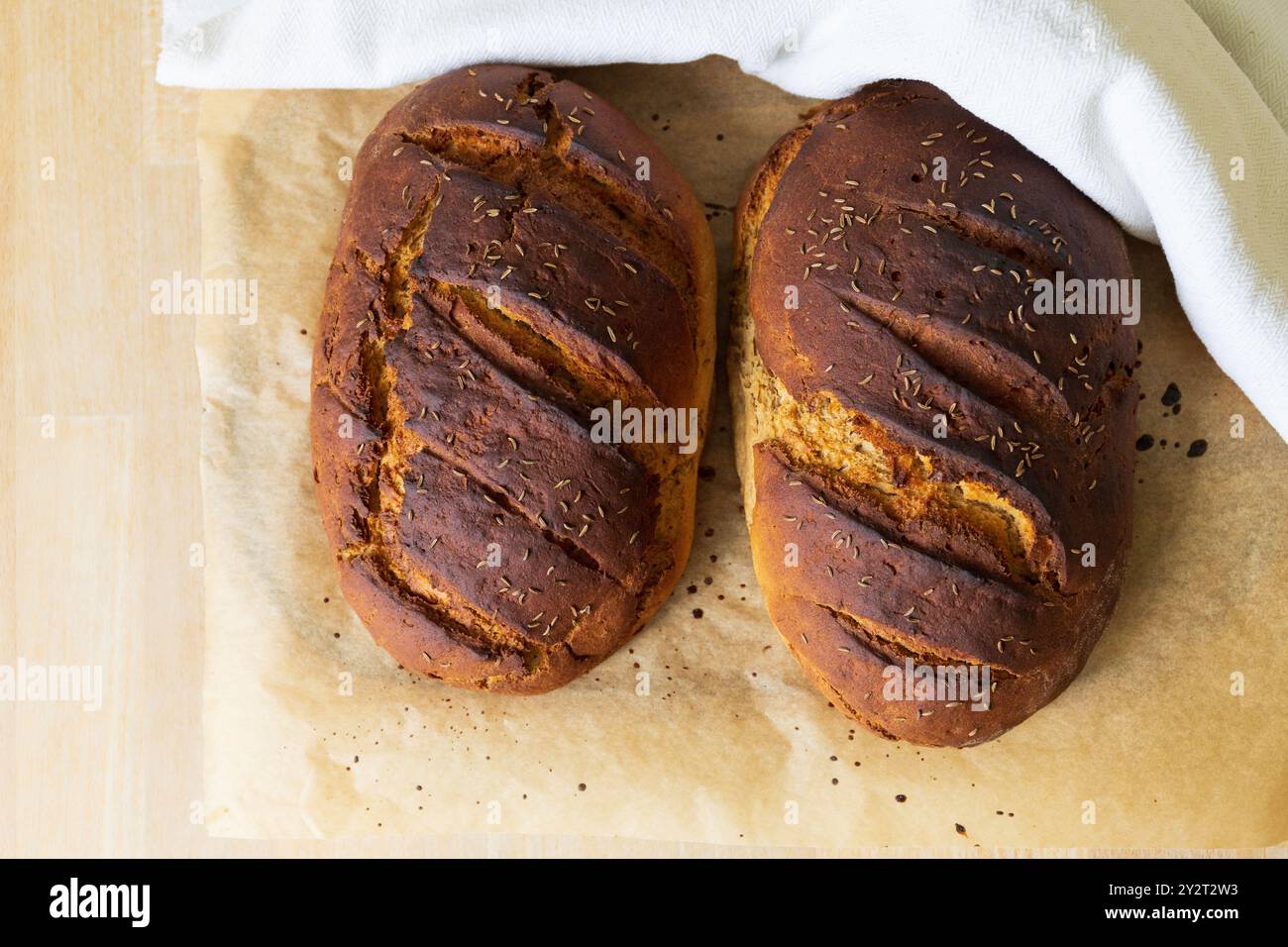 Photographie de table de deux pains de seigle fraîchement cuits sur un papier cuisson partiellement recouvert d'un torchon blanc à la lumière du jour Banque D'Images