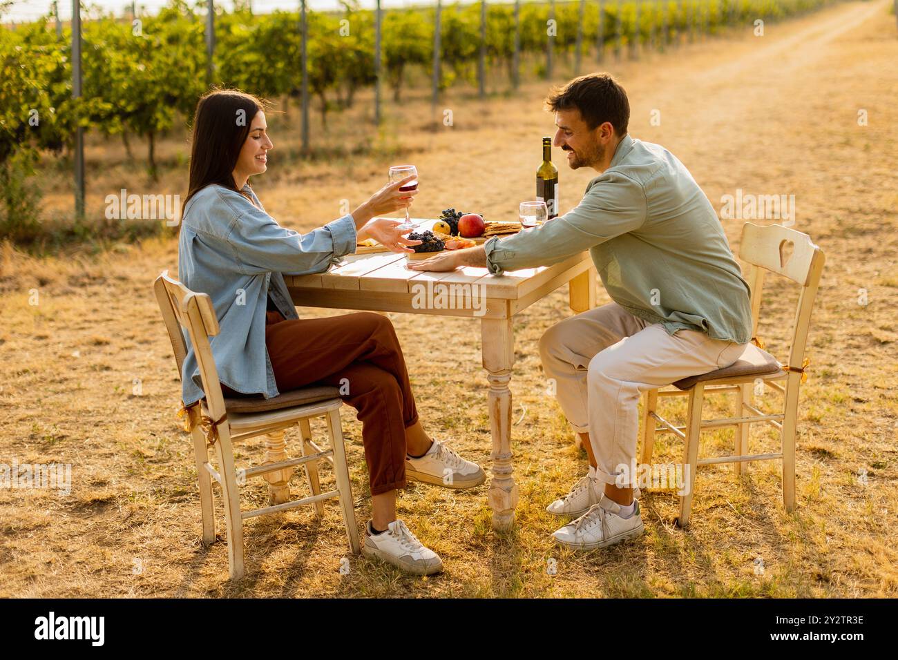 Couple se détend à une table rustique dans un vignoble ensoleillé, en savourant du vin et des collations gastronomiques. Ils partagent le rire et la joie, entourés de vignes luxuriantes et le Banque D'Images