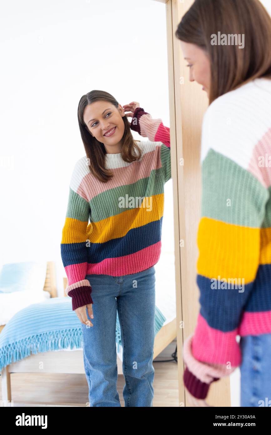 À la maison, femme souriante en pull coloré se regardant dans le miroir, se sentant heureuse Banque D'Images