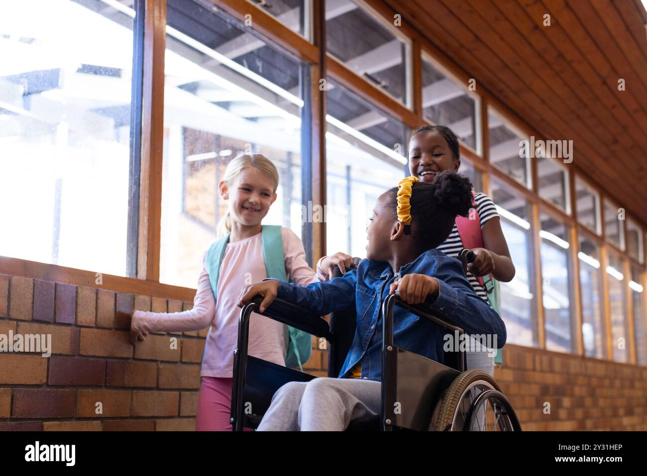 A l'école, trois filles multiraciales interagissent dans le couloir, une en fauteuil roulant, souriant ensemble Banque D'Images