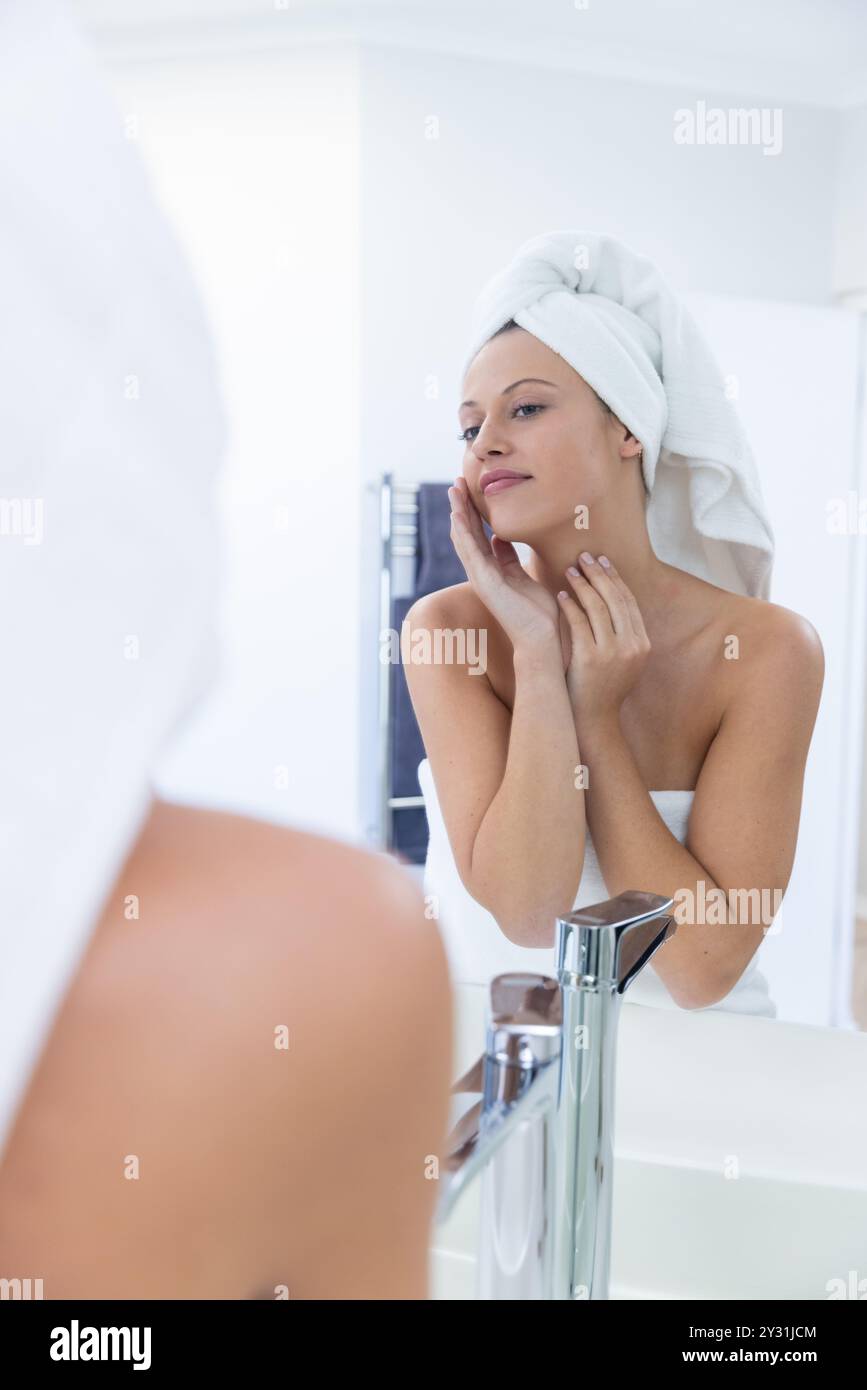 À la maison, en appliquant des soins de la peau, jeune femme avec une serviette sur la tête regardant dans le miroir de la salle de bain Banque D'Images