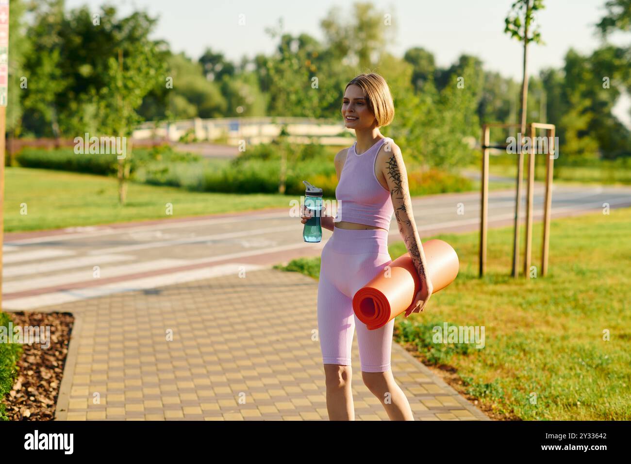 Une jeune femme joyeuse marche dans un parc, tenant un tapis de yoga et une bouteille d'eau. Banque D'Images