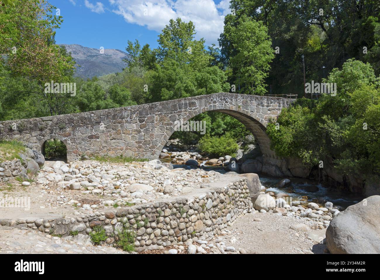 Un pont de pierre historique traverse un lit de rivière dans un paysage montagneux et boisé sous un ciel bleu, Puente Parral, pont romain Parral, Garganta Jaranda, Banque D'Images