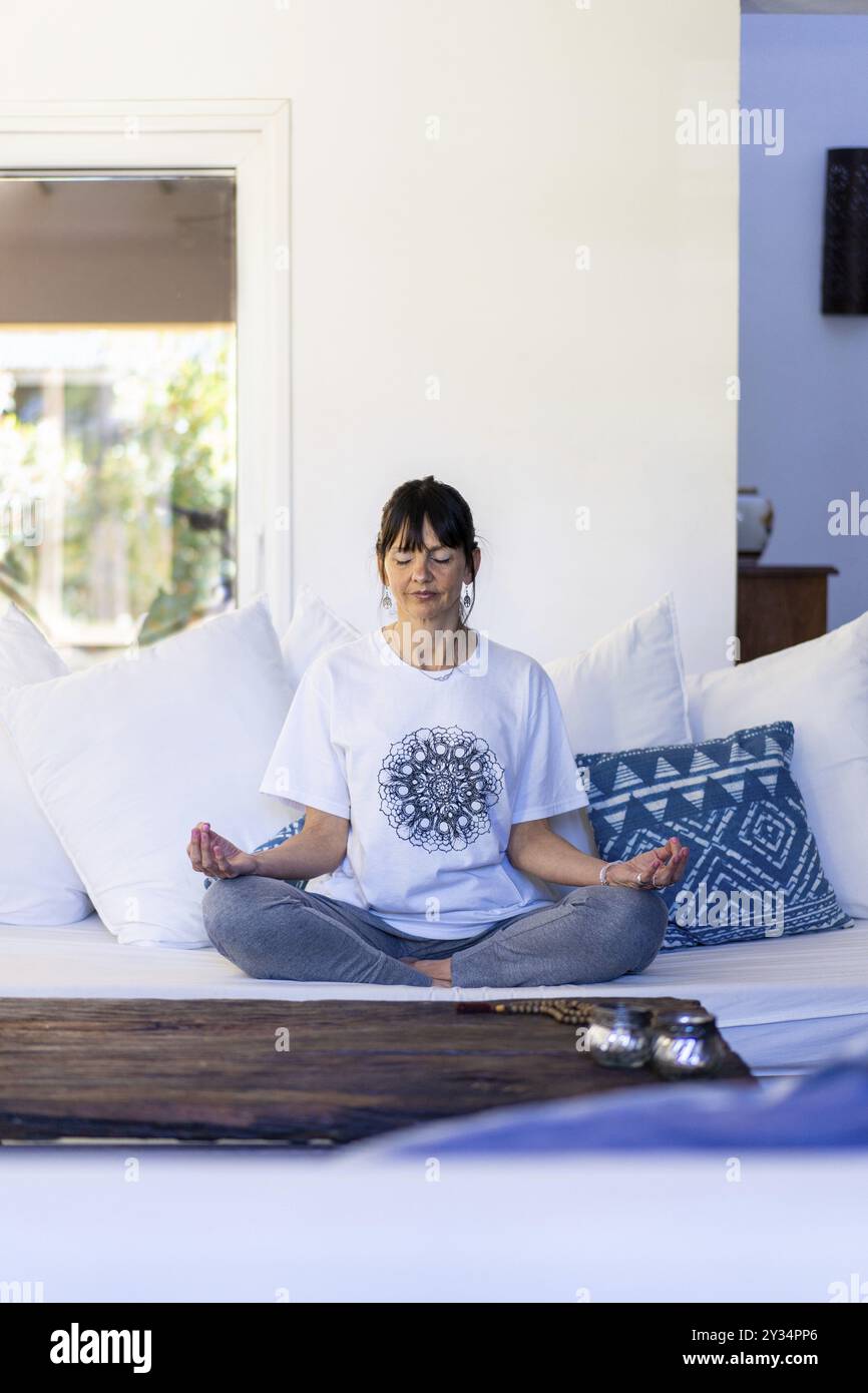 Femme mi-adulte pratiquant la méditation sur son canapé pendant la journée, éclairée par la lumière naturelle, avec la frange et ses cheveux attachés, les mains en Gyan Mudra Banque D'Images