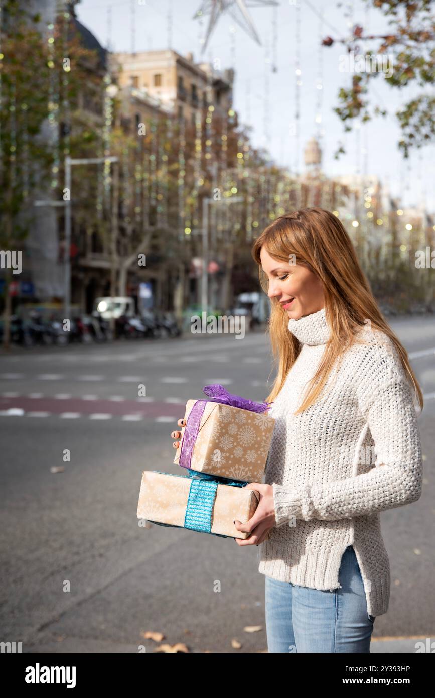 Femme blonde souriante avec des cadeaux de Noël dans une rue de Barcelone, ornée de lumières festives, avec un espace pour la copie. Banque D'Images