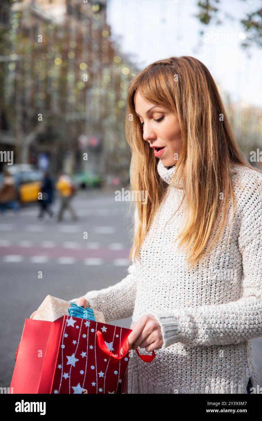 Femme surprise tirant un cadeau d'un sac étoilé rouge sur une rue décorée de façon festive de Barcelone, avec un espace vertical pour le texte. Banque D'Images