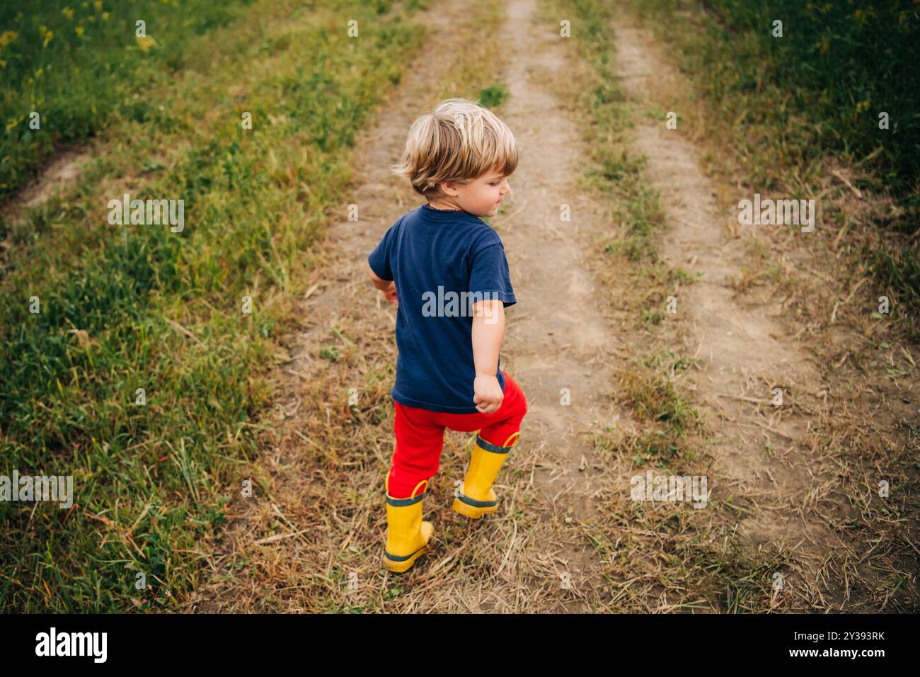 Un enfant marche sur un chemin de terre dans un champ, portant des bottes jaunes Banque D'Images