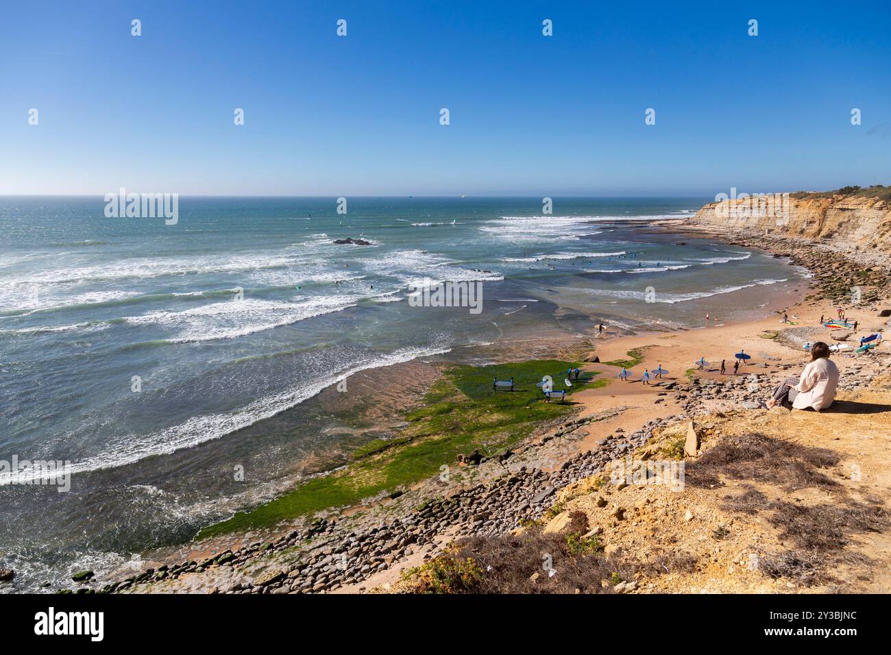 Femme assise au sommet d'une falaise donnant sur l'océan Atlantique et Matadouro Beach à Ericeira à marée basse par une journée venteuse. Portugal Banque D'Images