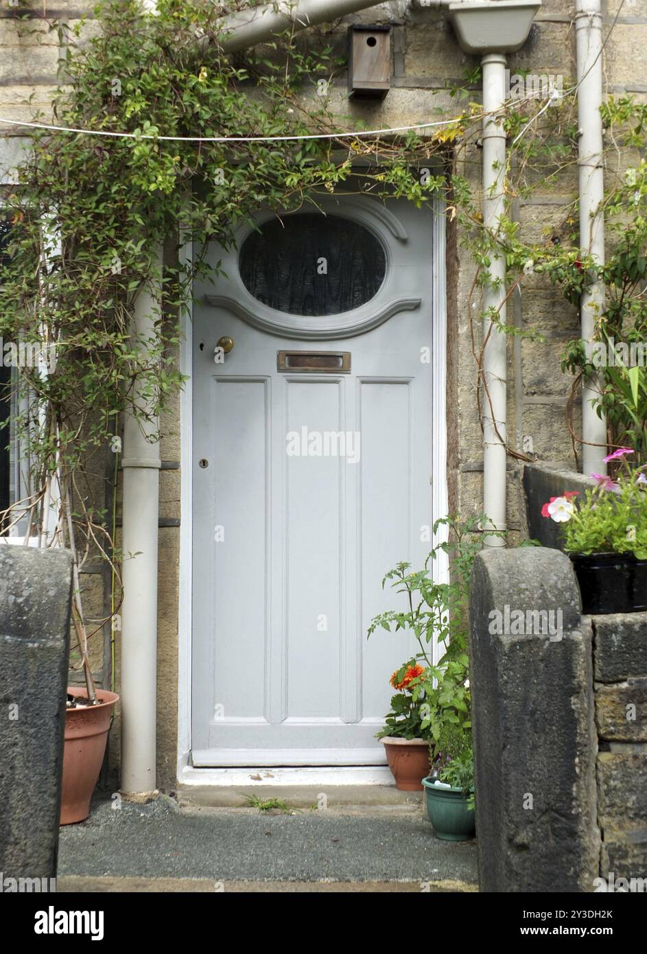 Porte de maison traditionnelle en bois peint en gris bleu avec boîte aux lettres en laiton et fenêtre avec un cadre blanc entouré de fleurs, de plantes en pot et d'un wal en pierre Banque D'Images