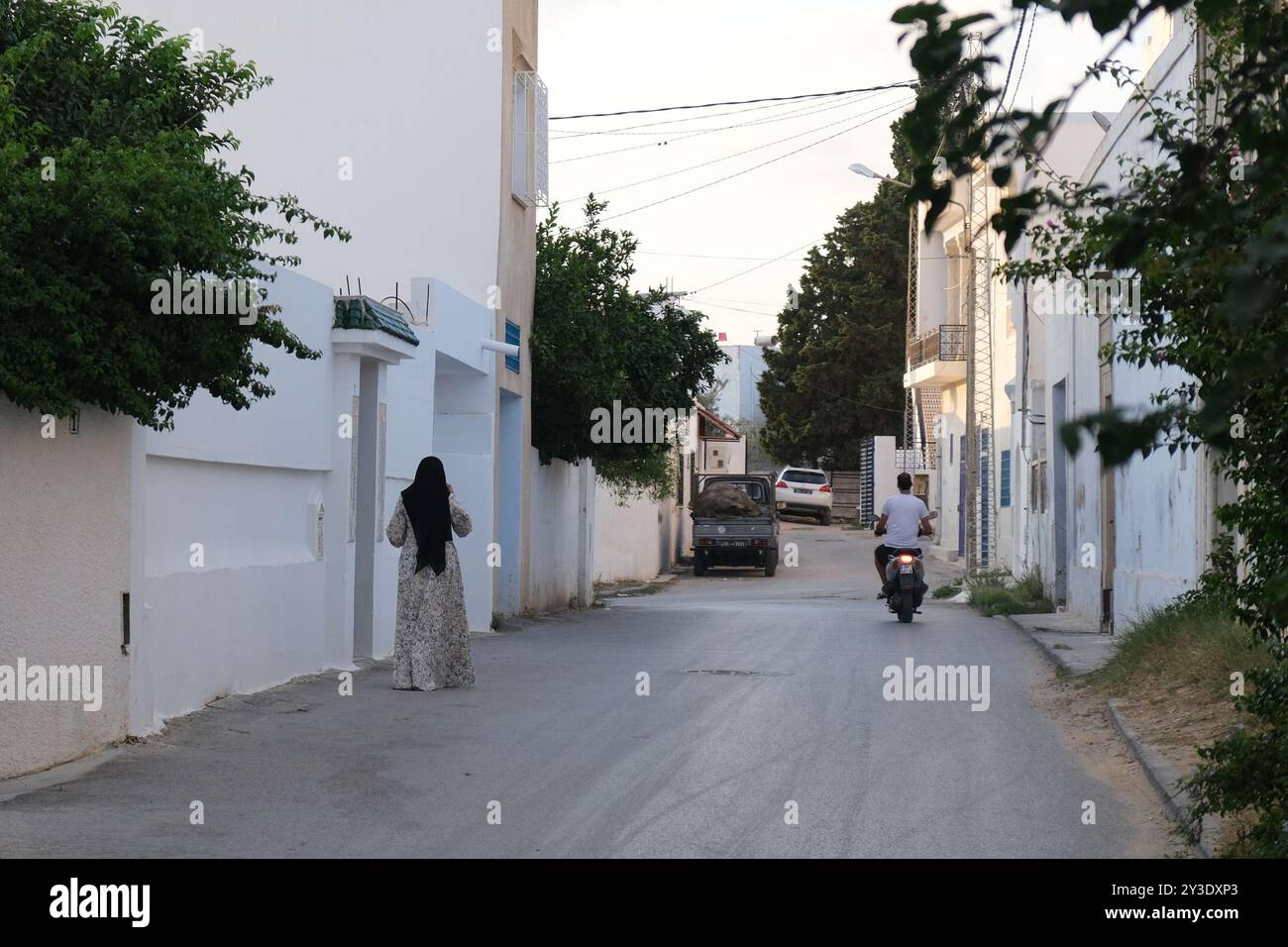 Une femme se tient debout comme un homme qui monte un scooter dans une rue latérale de la ville tunisienne de Hammamet dans le soleil de la fin de l'après-midi Banque D'Images