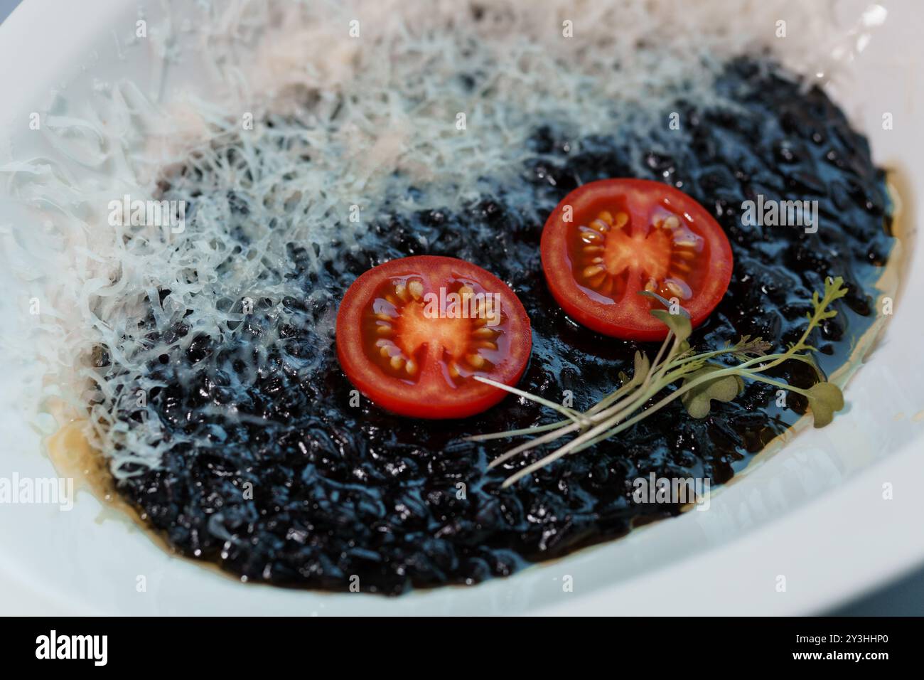 Risotto noir gourmand avec fromage râpé et tomates cerises fraîches garnies de Microgreens sur une élégante assiette blanche. Banque D'Images