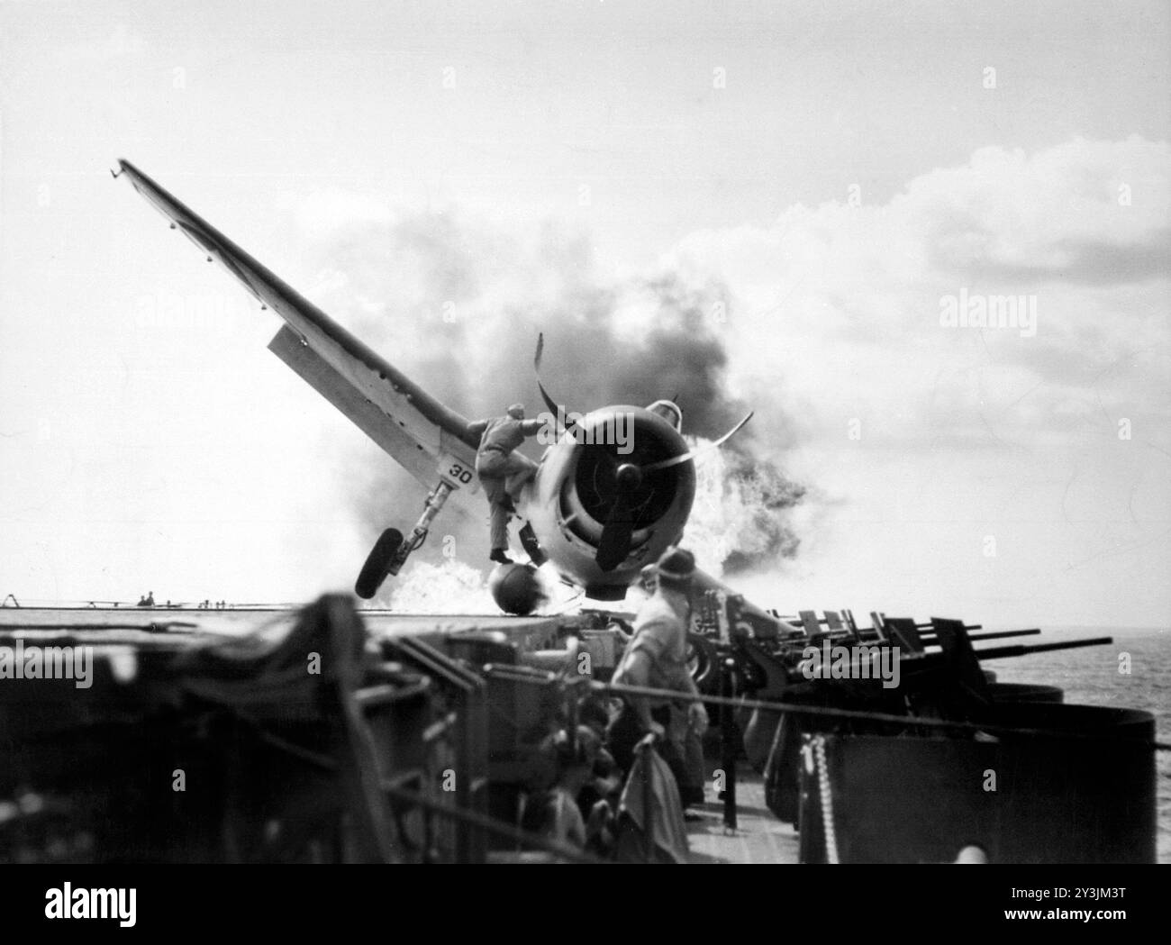 Atterrissage écrasé d'un Grumman F6F-3 Hellcat (numéro 30) du Fighting Squadron 2 (VF-2) à bord du porte-avions USS Enterprise (CV-6), dans la galerie de canons de 20 mm côté bâbord du porte-avions, le 10 novembre 1943. Le lieutenant Walter L. Chewning, Jr., USNR, l'officier de catapulte, est vu grimper sur le côté de l'avion pour aider le pilote à échapper à l'avion en feu. Le pilote, l'enseigne Byron M. Johnson, s'est échappé sans blessure grave. L'Enterprise était alors en route pour soutenir l'opération Gilberts. Notez le réservoir de carburant ventral rompu de l'avion. Photo de l'US Navy. Banque D'Images