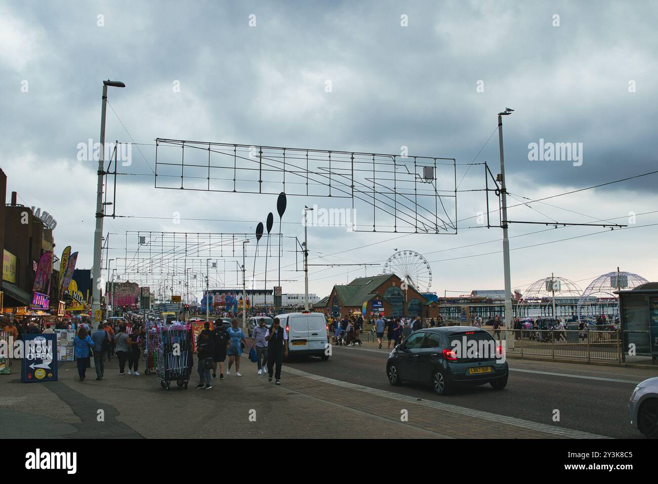 Une promenade balnéaire animée avec des gens marchant, des boutiques sur le côté, et un ciel nuageux. Il y a des manèges en arrière-plan, y compris un Ferris whe Banque D'Images