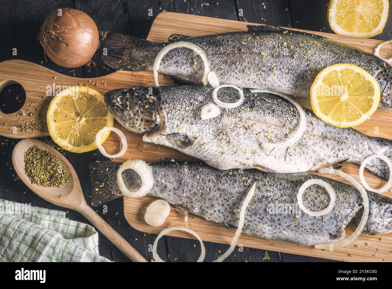 Gros plan avec de la truite fraîche, saupoudrée d'herbes, d'épices, de rondelles d'oignon et de tranches de citron, placées sur une planche à découper en bois, sur une table vintage Banque D'Images