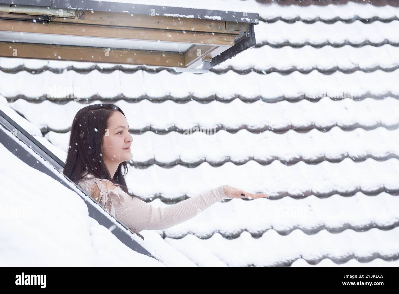 Jolie femme brune vêtue d'un pull confortable debout à l'extérieur sur sa fenêtre atteignant sa main pour attraper les flocons de neige Banque D'Images