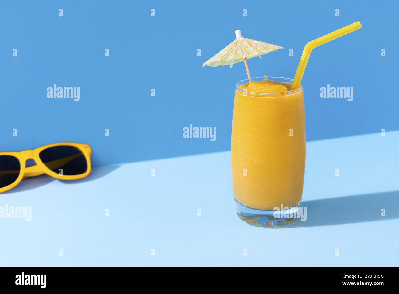 Verre de smoothie à la mangue et lunettes de soleil jaunes sur un fond bleu, en forte lumière. Contexte ensoleillé de la journée d'été. Smoothie tropical. Cocktail à la mangue Banque D'Images