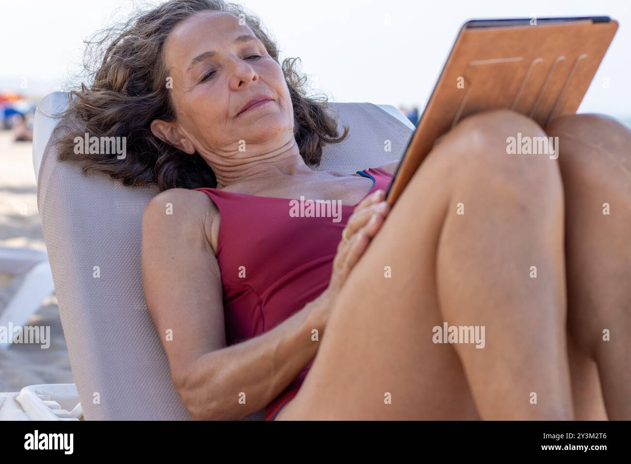 Une femme âgée est confortablement allongée sur une chaise de plage, lisant ou regardant une tablette. Banque D'Images