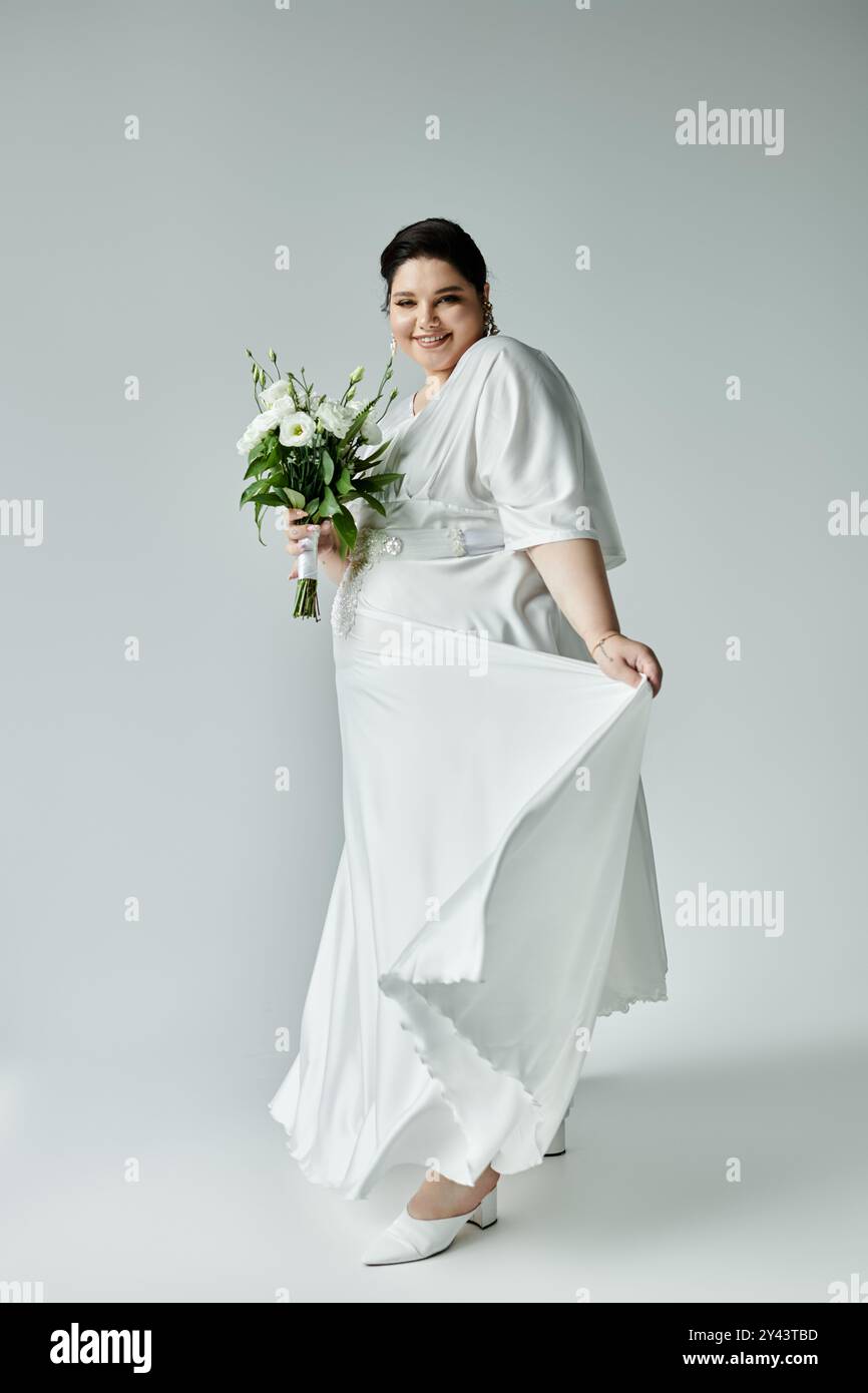 Une joyeuse mariée grande taille dans une superbe robe blanche tient gracieusement son bouquet sur un fond serein. Banque D'Images