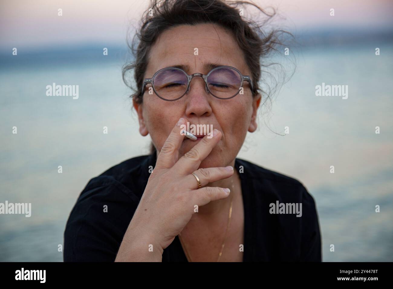 Femme d'âge moyen avec des lunettes fumant sur la plage au coucher du soleil Banque D'Images