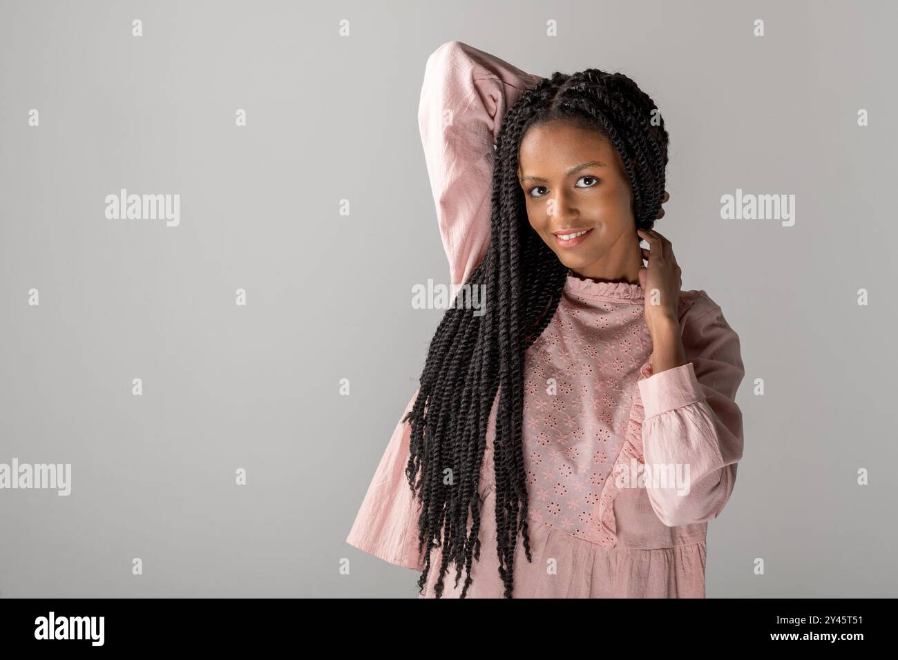 Portrait de jeune femme afro-américaine souriante avec de longues tresses regardant la caméra tout en se tenant debout avec la main levée derrière la tête contre la grotte grise Banque D'Images